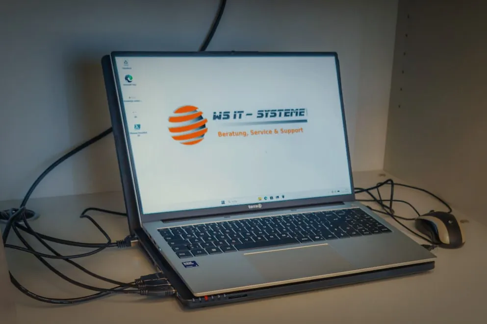 Notebook mit dem Logo von WS IT-Systeme im Fach eines Büroschranks, daneben Maus und Kabel.