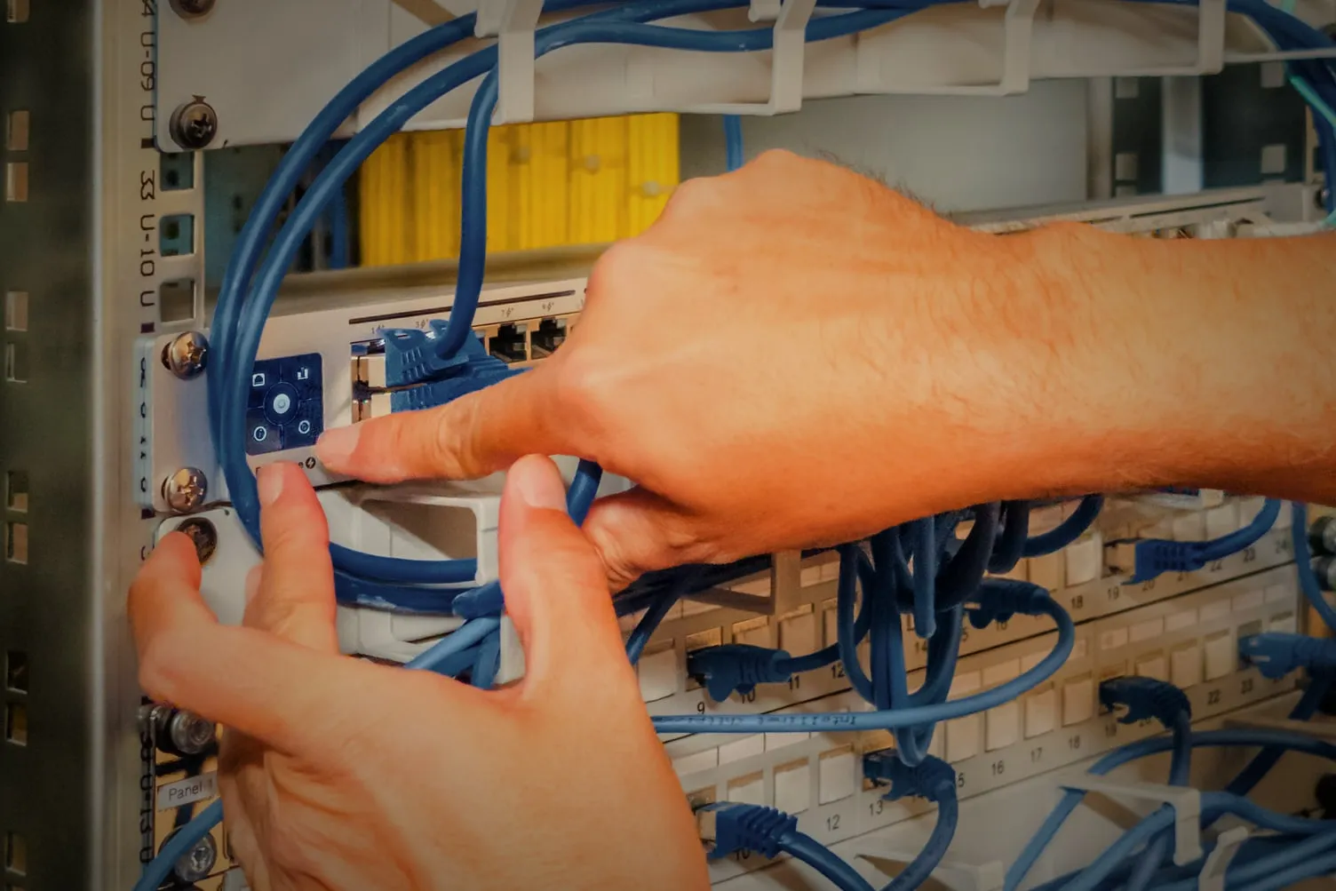 Hände stecken blaue Patchkabel an Switch und Patchfeld im Netzwerkschrank.